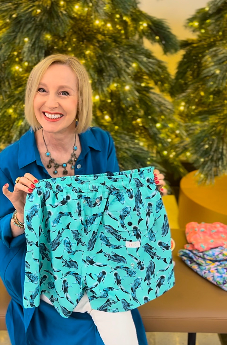 Ann Vodicka, Sydney based personal colour analyst, is smiling and holding a pair of aqua and navy blue whale patterned boardshorts.